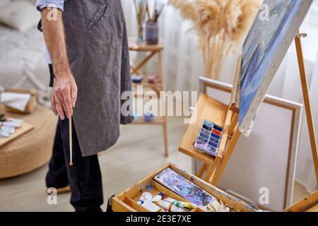 uomo pittore maschile in grembiule durante il lavoro, maschio con pennelli, materiali vari vernici strumenti per la pittura Foto Stock