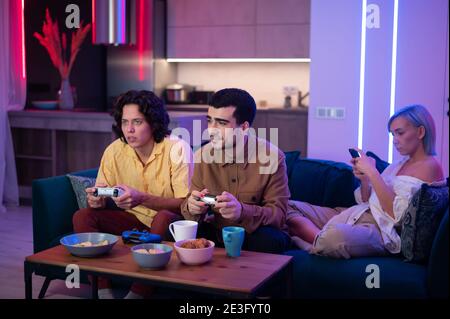 La compagnia degli amici si rilassa sul divano giocando a videogiochi e divertendosi in un moderno appartamento illuminato con colore neon. Foto Stock