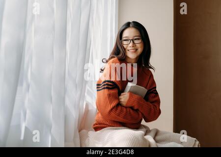 Donna asiatica felice in occhiali e maglione hugging libro e. guardando via con il sorriso mentre si riposa vicino alla finestra di casa Foto Stock