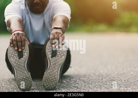 Primo piano Asian giovane atleta sportivo corridore nero uomo indossare orologio egli seduta tirare piedi allungare gambe e ginocchio prima di correre a strada guarire all'aperto Foto Stock
