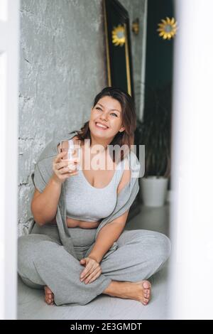 Bella bruna felice giovane donna più corpo taglia positivo in comodo abbigliamento sportivo con bicchiere d'acqua a casa Foto Stock