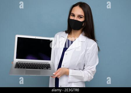 Giovane donna di brunetto che indossa camice medico bianco e maschera nera tenendo il computer portatile e guardando la fotocamera isolata sulla parete blu sfondo Foto Stock