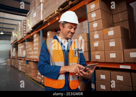 Lavoratore maschile felice che lavora in fabbrica utilizzando il supporto di un tablet digitale vicino alla pila di scatole di cartone Foto Stock