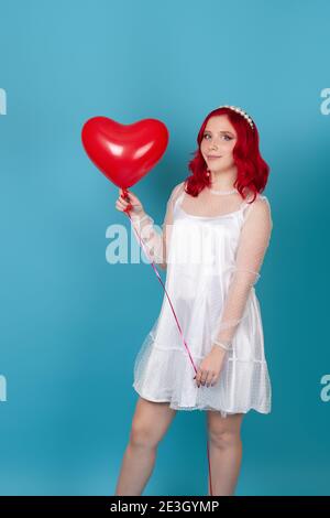 giovane donna carina in abito bianco e con capelli rossi tenendo un pallone volante a forma di cuore isolato su uno sfondo blu Foto Stock