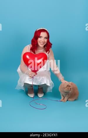 Una giovane donna sorridente in un vestito bianco tiene un pallone volante a forma di cuore, sbatte e spreme un gatto britannico rosso, isolato su uno sfondo blu Foto Stock