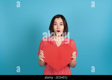 primo piano sorpresa, eccitato, impressionato giovane donna asiatica con bocca aperta in abito rosso tenendo grande cuore di carta rosso isolato su sfondo blu Foto Stock