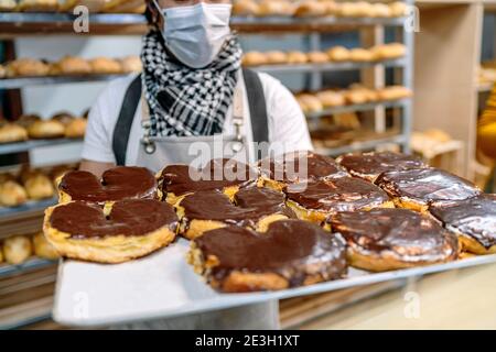 Il panettiere ha un vassoio di cioccolato da forno palme fresco dal forno e indossa una maschera per il viso a causa di il 2020 covid19 coronavirus pandemia Foto Stock