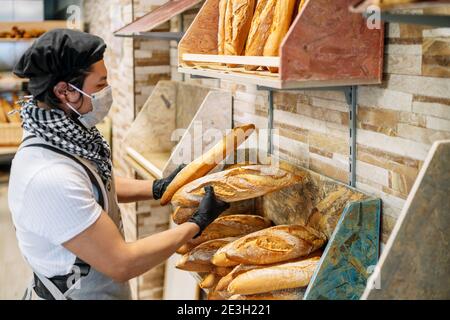 Fuoco selettivo di panettiere latino con scaffalatura pane appena sfornato una maschera protettiva per il coronavirus 2020 covid 19 pandemia Foto Stock