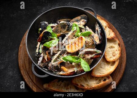 Cozze cotte in padella con formaggio e foglie di basilico, su tavola di legno, su fondo scuro Foto Stock