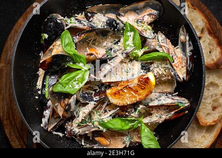 Cozze cotte in padella con formaggio e foglie di basilico, su tavola di legno, su fondo scuro Foto Stock