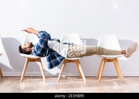 Giovane uomo che usa lo smartphone mentre si sdraiò sulle sedie nella sala Foto Stock