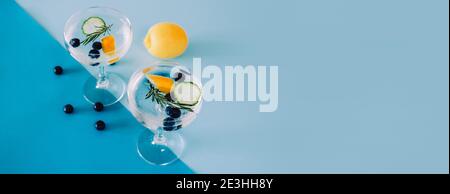 Banner con due bicchieri di Gin e cocktail tonici con cetrioli, limone e mirtilli isolati su sfondo blu geometrico Foto Stock