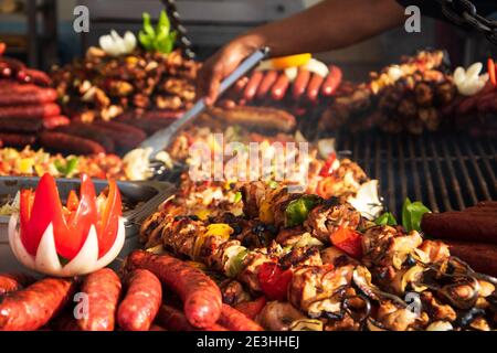 Salsicce e kebab grigliati assortiti su un grande grill rotondo al mercato Street food di Parigi, Francia. Fornitore che sceglie salsicce per preparare il sandwich. Foto Stock