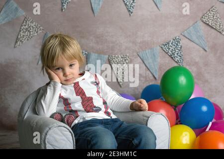 Carino bambino biondo, ragazzo, seduto su piccola poltrona bambino, giocando con palloncini colorati, sorridendo felicemente, tiro al coperto Foto Stock