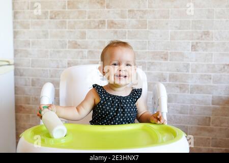 Bambino felice, ragazza del toddler, godendo la colazione sana e bevendo il latte seduto in sedia alta alla cucina soleggiata luminosa Foto Stock