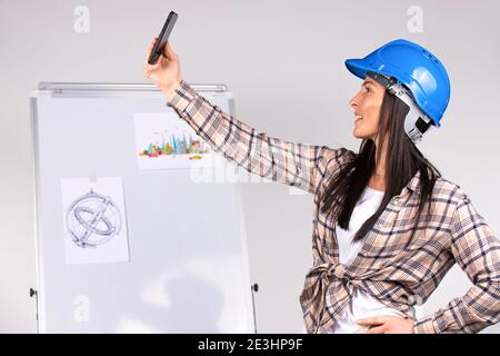 Giovane ingegnere femmina sorride e fa selfie su smartphone ufficio sfondo grigio e diagrammi. Foto Stock