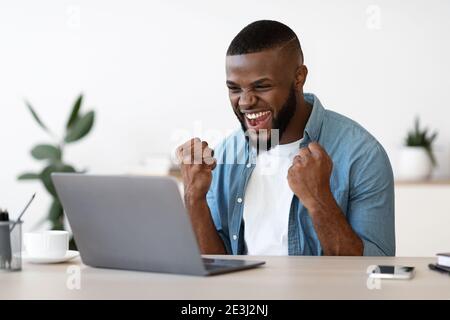 Gioioso Black Guy con i notebook Raising Fists sul luogo di lavoro, celebrando il successo aziendale Foto Stock