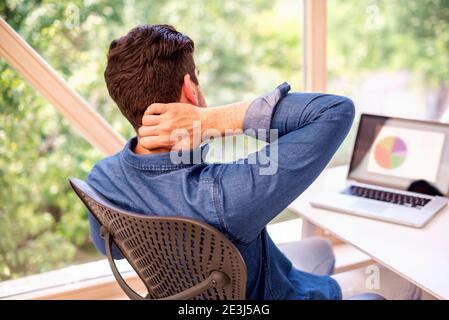 Vista posteriore di un uomo d'affari irriconoscibile con la mano sul tempio mentre si siede dietro il suo laptop e soffre di forte dolore alla testa. Foto Stock