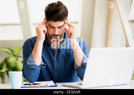 Scatto di un uomo d'affari con gli occhi chiusi e le mani sul tempio, mentre si siede dietro il suo laptop e soffre di forte dolore alla testa. Foto Stock