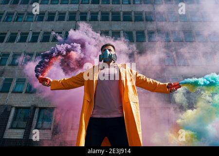 Il giovane uomo in maschera respiratore regge bombe di fumo in mani alzate. Protesta, libertà, concetto di rivoluzione Foto Stock