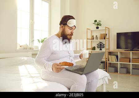 Uomo divertente in pigiami bianchi seduti con caffè e croissant a letto e guardando il computer portatile Foto Stock