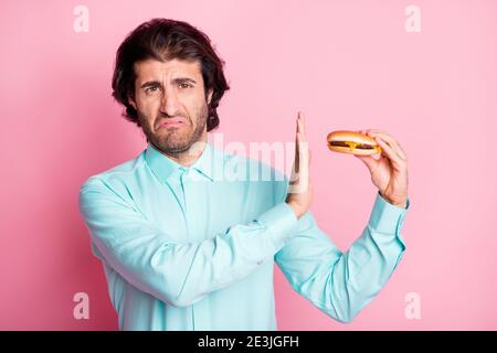 Ritratto fotografico di uomo disgustato tenendo cheeseburger in mano rifiutando con palma isolata su sfondo color rosa pastello Foto Stock