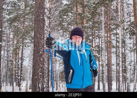 Ritratto di un uomo anziano felice in una giacca blu e un cappello nero lavorato a maglia, sci di fondo sullo sfondo di una foresta innevata invernale. Chiudi-u Foto Stock