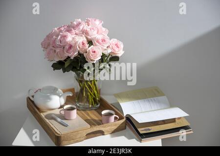 Rosa Bianco Rosa o'hara. Bouquet di rose rosa in vaso di vetro con una teiera bianca e due tazze di cappuccino su un vassoio su un tavolino tra i libri. Foto Stock