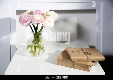 Un bouquet di bicchierini rosa persiani in un vaso di vetro e due regali avvolti su un tavolo bianco di fronte ad un falso camino bianco. Interno. Foto Stock