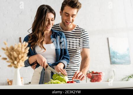 felice coppia preparare insalata in cucina con catkins su sfocato primo piano Foto Stock