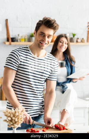 uomo sorridente vicino a donna tenendo calice e giornale su sfocato sfondo Foto Stock