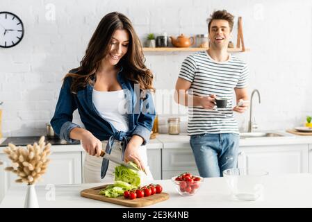 donna allegra che cucina mentre l'uomo tiene la tazza e lo smartphone sopra sfondo sfocato Foto Stock