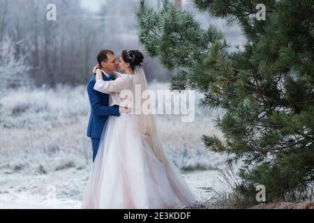 sposi in una passeggiata di nozze in inverno stand insieme e posa per un fotografo vicino a un albero coperto di neve in una bella foresta, la famiglia scatta una foto Foto Stock