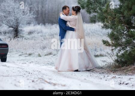 sposi in una passeggiata di nozze in inverno stand insieme e posa per un fotografo vicino a un albero coperto di neve in una bella foresta, la famiglia scatta una foto Foto Stock