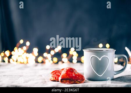 Biscotti rossi a forma di cuore fatti a mano e tazza di caffè grigia e luci. Concetto di San Valentino. Foto Stock