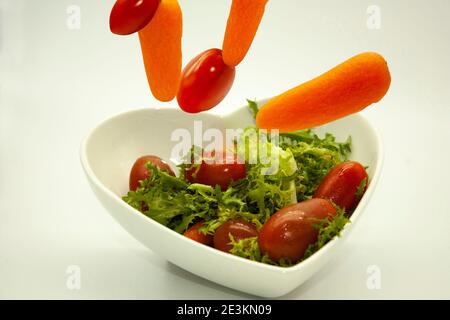 Verdure che cadono in ciotola a forma di cuore con insalata vegana organica grezza combinazione di lecguga, pomodori ciliegia rossa, e carote Foto Stock