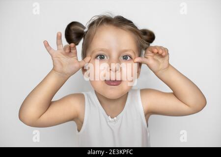 Primo piano di studio di cute adorabile bambina, indossando top bianco, con due ponytail, rendendo divertente viso e mostrare lingua. Ragazzina che stuzzicava Foto Stock