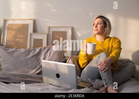 Una donna si siede su un letto e lavora con un computer portatile e una tazza di tè e guarda fuori Finestra durante il Coronavirus Covid-19. Il concetto di speranza per un fine Foto Stock