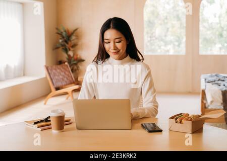 Giovane donna d'affari asiatica con occhiali e computer portatile sorridente guardando la macchina fotografica. Donna freelance seduto al tavolo e che lavora da casa. Foto Stock