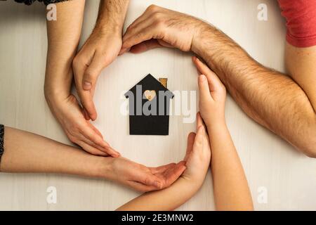 Mani di genitori e bambini circondano il layout di una casa scura su uno sfondo chiaro. Famiglia, immobili e assicurazioni, appartamento, vista dall'alto. Foto Stock