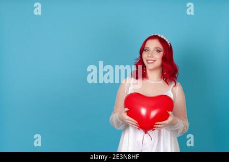 ridendo gioiosa giovane donna in un vestito bianco con rosso i capelli trattengono un pallone volante a forma di cuore isolato su un blu sfondo con entrambe le mani Foto Stock