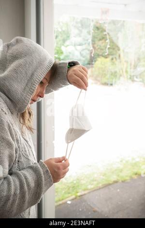 Una donna di mezza età si trova alla finestra guardando una maschera FFP 2. È preoccupata per l'incerto futuro della società a causa della pandemia di Corona. No Foto Stock