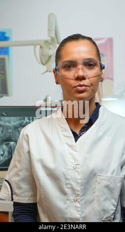 Medico dentista di successo che ha un discorso sui denti igienici, guardando la fotocamera, parlare fiducioso in clinica dentale mentre assistente che prepara l'uomo per la chirurgia stomatologica in background. Foto Stock