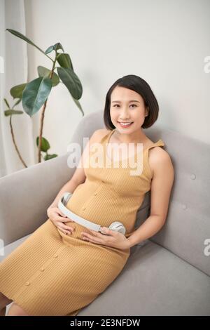 Bella donna incinta che mette le cuffie sul ventre mentre si siede sul divano Foto Stock