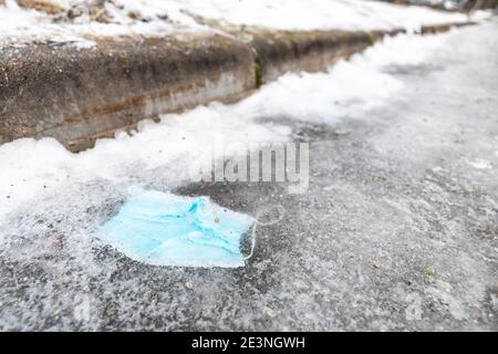 Maschera usata congelata in ghiaccio sulla strada. Le maschere chirurgiche blu chiaro cadono sul marciapiede. Arrestare l'inquinamento Foto Stock