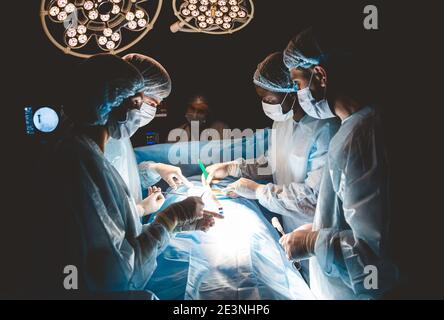 Un team professionale internazionale di chirurgo, assistenti e anestesista esegue un'operazione complessa su un paziente sottoposto ad anestesia generale. Scuro Foto Stock