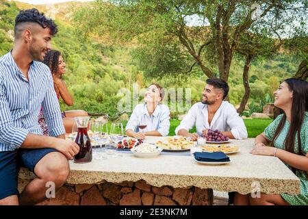 Gruppo di giovani di razza mista che si divertono all'aperto in campagna mangiando spuntini e bevendo vino rosso per un aperitivo. Bella amici felici bo Foto Stock