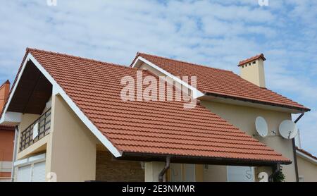 Un primo piano di stucco casa con tetto a doppio tetto in tegole di metallo rosso, un camino, pannelli di plancia, grondaie a pioggia, due antenne paraboliche satellitari e un attico. Foto Stock