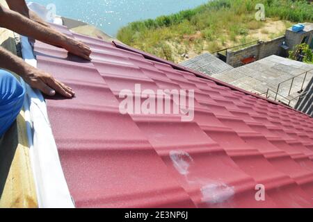 Costruzione di tetti. Un appaltatore dell'edificio sta installando tegole del tetto corrugate in metallo rosso, scudi e rivestimenti dell'alata sulla cresta del tetto. Foto Stock