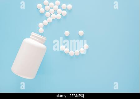 faccia sorridente fiancheggiata da pillole. Vasetto bianco e pillole bianche sparse. Faccia sorridente fatta da pillole rotonde bianche su sfondo blu. Medicina e salute c Foto Stock